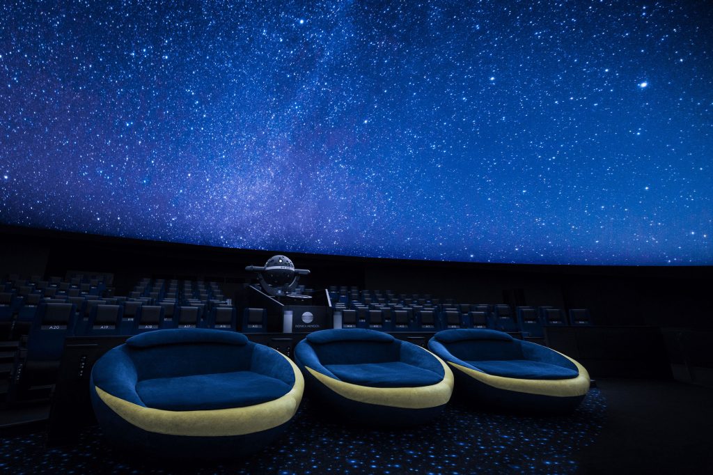 コニカミノルタプラネタリウム天空のシアタールームの写真。満点の空が写っているスクリーンとその下にはシートと映写機。手前に3台寝転んで星空を見ることのできる三日月シート。